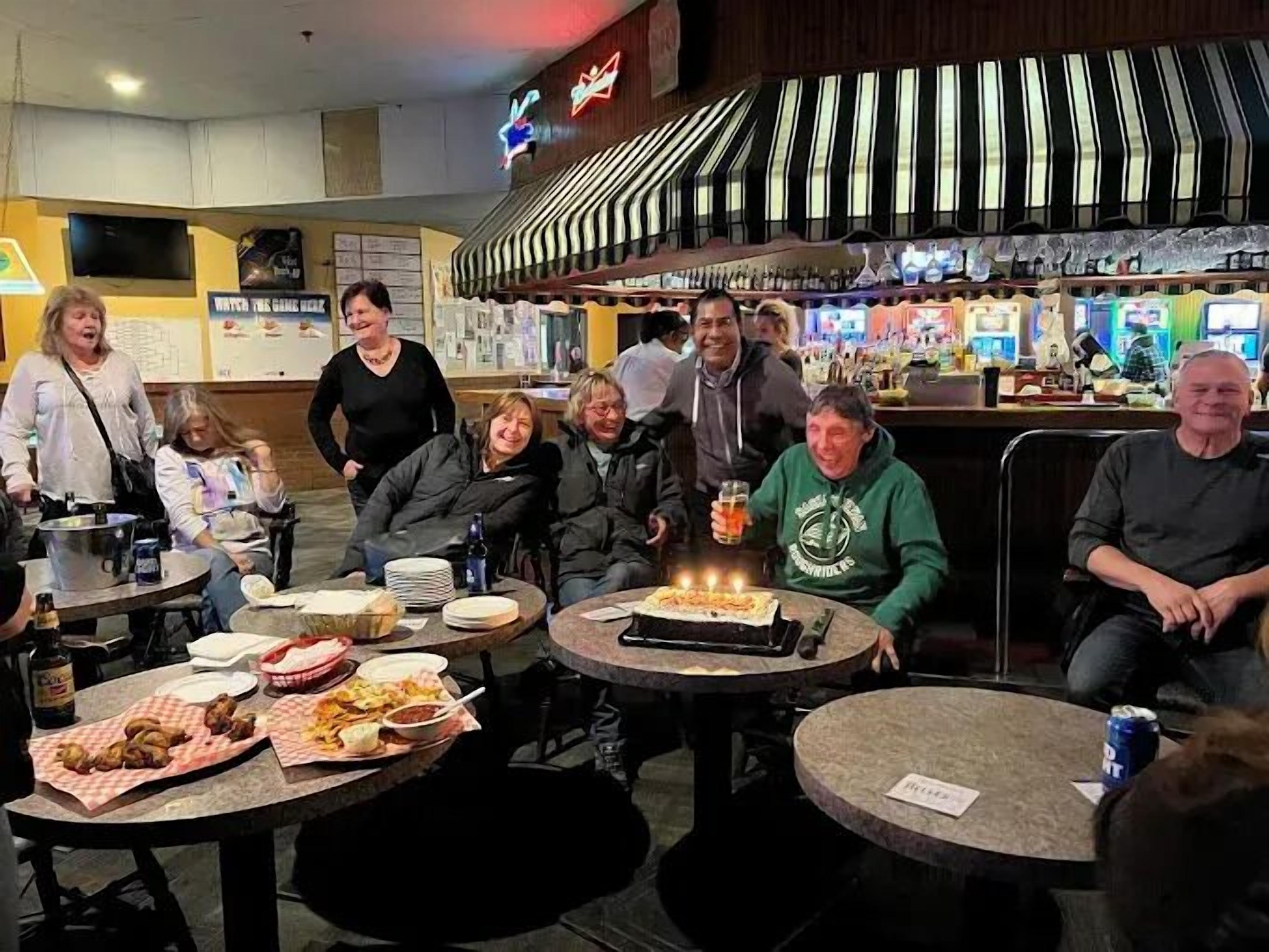 A group of people seated around tables with food and drinks, in a restaurant with a bar and counter visible in the background.