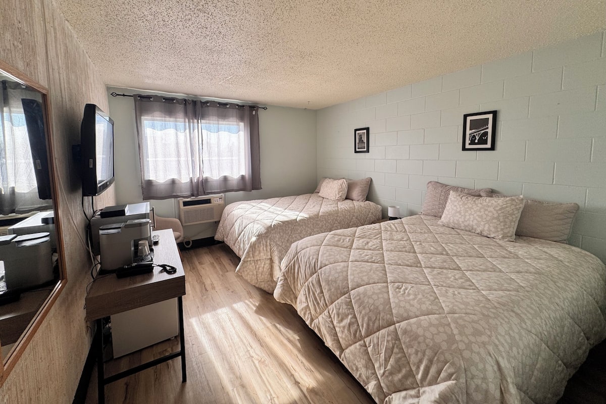 A room with two beds, a desk with a computer, a TV, and a window with curtains.