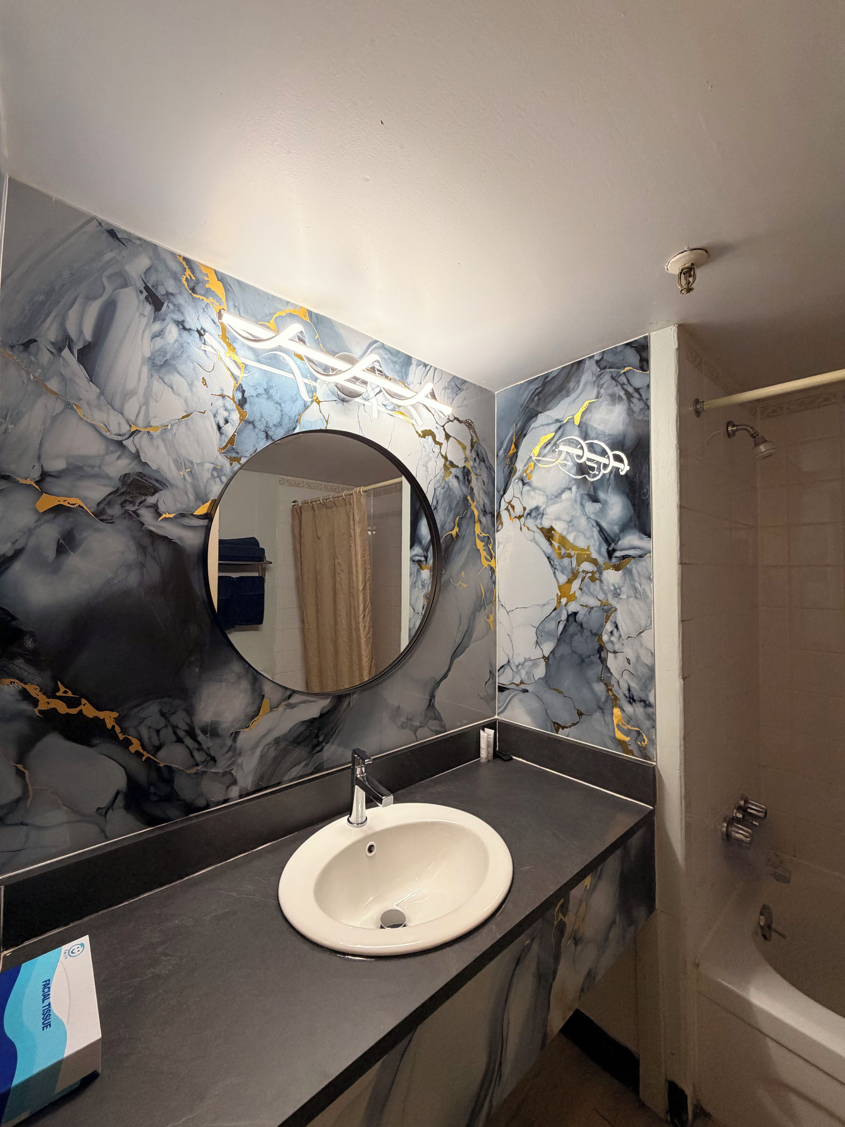 Een badkamer met een marmeren wand, een ronde spiegel, een wastafel en een douche.