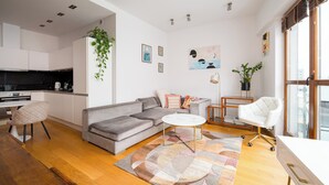 A modern living room with a grey sofa, a round coffee table, and a white armchair. There is a kitchen area with white cabinets and a black countertop.