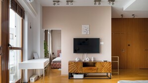 A modern living room with a white desk, a flat-screen TV mounted on a wooden cabinet, a large window with blinds, and a wooden door.