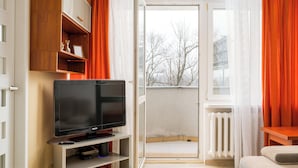 A modern apartment with a TV on a stand, wooden cabinets, a balcony with orange curtains, and a view of trees. - Solec Powiśle Harmony Apartment (Warszawa)