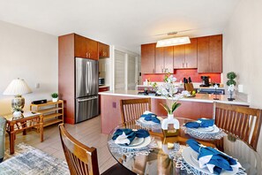 A modern kitchen with wooden cabinets, a stainless steel refrigerator, and a dining area with a glass table set for four. - New Listing! South Maui Condo Steps to Beach (Kihei)