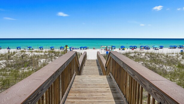 Una passerella in legno conduce a una spiaggia con ombrelloni blu e cieli sereni.