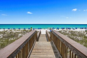 Ein Holzsteg führt zu einem Strand mit blauen Sonnenschirmen und blauem Himmel.