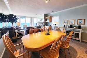 A spacious living room with a large wooden dining table surrounded by chairs, a cozy seating area with a sofa and armchairs, a kitchen area with a countertop and cabinets, and a view of the ocean through large windows.