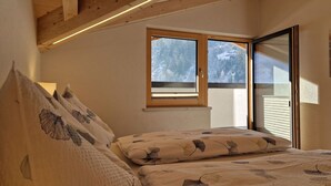 Une chambre avec un plafond en pente, des poutres en bois, un lit recouvert d’une literie à motifs floraux et une fenêtre donnant sur des arbres enneigés.