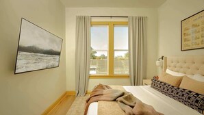 Ein Schlafzimmer mit einem großen Bett, einem Fenster mit Vorhängen, einem Gemälde an der Wand und einem Kopfteil mit einer Leuchte.