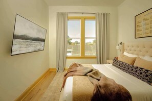Een slaapkamer met een groot bed, een raam met gordijnen, een schilderij aan de muur en een hoofdeinde met een lamp.
