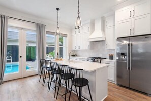A modern kitchen with white cabinets, a stainless steel refrigerator, and a bar area with stools. - Luxurious Marigny Home | Heated Pool (New Orleans)