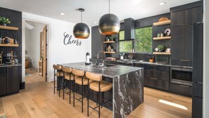 A modern kitchen with a marble island, bar stools, pendant lighting, and dark cabinetry. - Luxe Eagles Nest Home: 3-Tier Deck, Hot Tub, Fire Pit & Sunset Views! (Banner Elk)