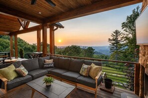 A covered outdoor seating area with a sofa and tables, overlooking a mountainous landscape. - Luxe Eagles Nest Home: 3-Tier Deck, Hot Tub, Fire Pit & Sunset Views! (Banner Elk)