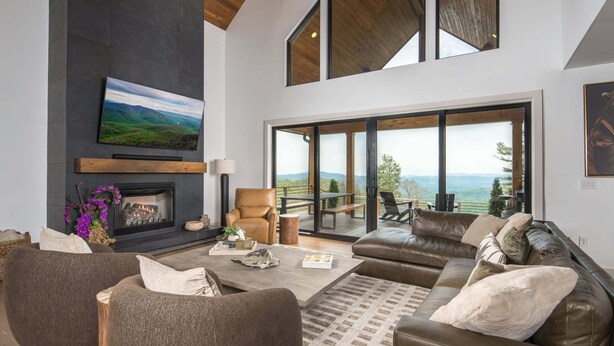 A modern living room with a fireplace, leather sofa, and a large window overlooking a scenic view. - Luxe Eagles Nest Home: 3-Tier Deck, Hot Tub, Fire Pit & Sunset Views! (Banner Elk)