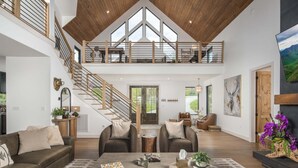 A spacious living area with a high ceiling, wooden staircase, and a large window. - Luxe Eagles Nest Home: 3-Tier Deck, Hot Tub, Fire Pit & Sunset Views! (Banner Elk)