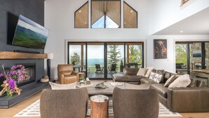 A spacious living room with a large sectional sofa, a fireplace, and a view of the outdoors through large windows. - Luxe Eagles Nest Home: 3-Tier Deck, Hot Tub, Fire Pit & Sunset Views! (Banner Elk)