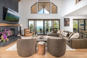 Ein geräumiges Wohnzimmer mit einem großen Ecksofa, einem Kamin und Blick ins Freie durch große Fenster.