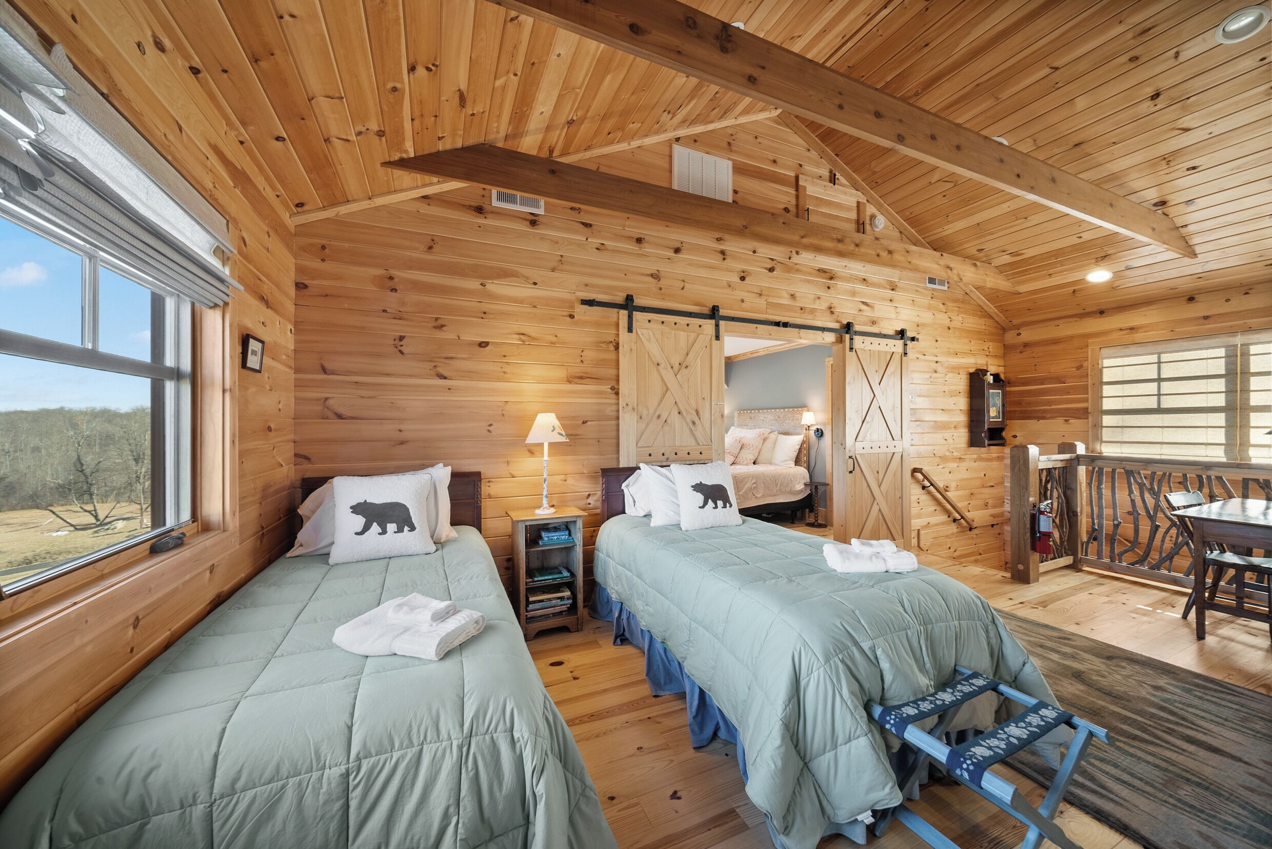A wooden cabin bedroom with two beds, a small table, a chair, and a window overlooking a forest.