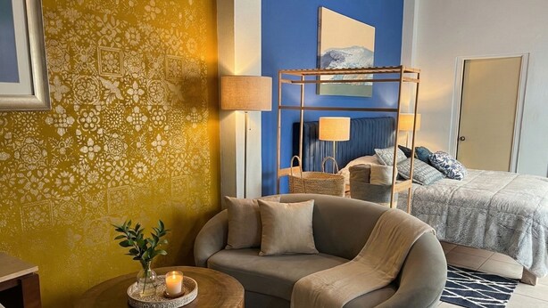 A cozy hotel room with a sofa, a small round wooden table with a candle, a bunk bed with a blue headboard, and a patterned yellow wall. - Casitas Aramara | Coastal Studio in Lo de Marcos (Lo de Marcos)