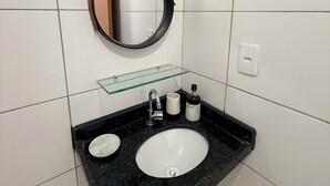 A bathroom with a black granite countertop, a white sink, a round mirror, and a glass shelf. - Comfortable and well-located apartment (João Pessoa)