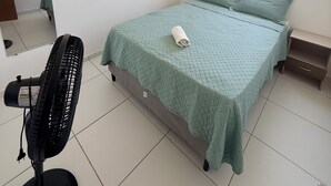 A bedroom with a bed, a standing fan, a window, and a shelf with a small item. - Comfortable and well-located apartment (João Pessoa)