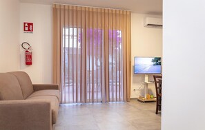 A living room with a beige sofa, a TV on a stand, a fire extinguisher, and a sliding door with a view of trees. - Lovely home in Santa Croce Camerina with house sea view (Santa Croce Camerina)