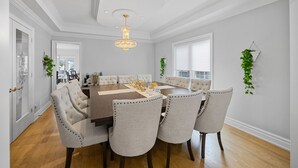 Une salle à manger avec une grande table en bois, six chaises rembourrées, un lustre et une vue sur l’extérieur grâce à de grandes fenêtres.