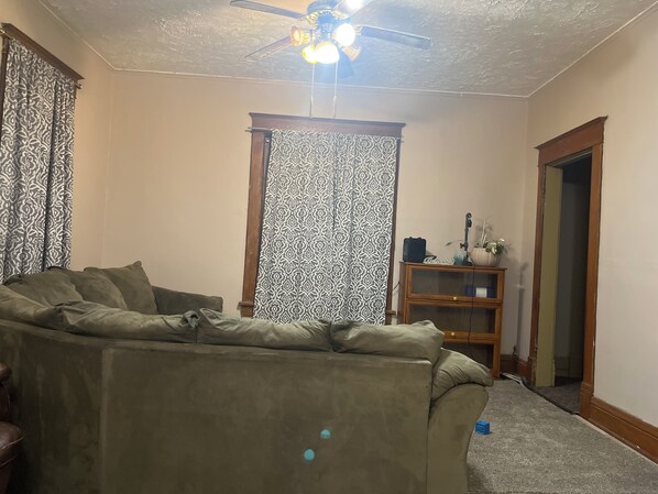 Una sala de estar con un sofá modular, un ventilador de techo, cortinas estampadas y un aparador de madera con adornos.