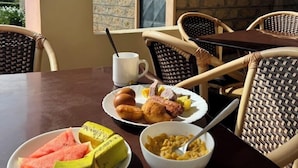 A dining area with a table set for breakfast, including plates of fruit, eggs, and cereal. There are wicker chairs and a view of a stone building in the background. - Alem Resort Tawa (Tawa)