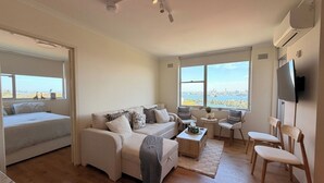 A spacious living area with a sofa, chairs, a coffee table, and a TV. There are large windows offering a view of a cityscape. - Mosman | Stunning Opera and Bridge View | KozyGuru (Mosman)