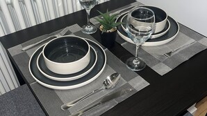 Ein Esstisch ist gedeckt mit schwarz-weißen Tellern, Besteck und klaren Gläsern. Im Hintergrund steht eine kleine Pflanze und ein Heizkörper.