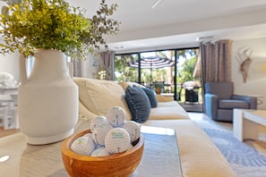 Ein Wohnzimmer mit einer Holtschale voller Golfbällen, einer weißen Vase mit Grünpflanzen, einer Couch mit Kissen und einem großen Fenster mit Blick ins Grüne.