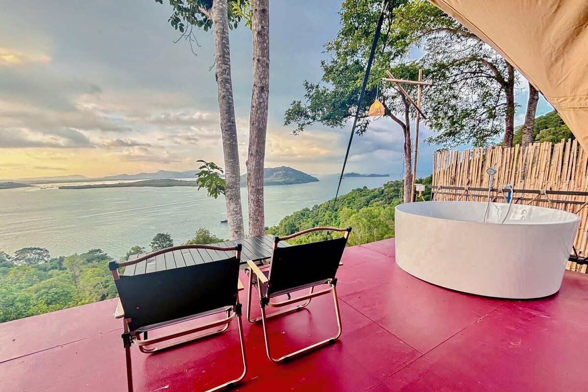 A red deck with two black chairs, a white tub, and a view of a lake and mountains.