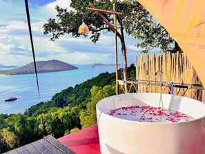 Un bain à remous rempli de pétales de rose, installé sur une terrasse offrant une vue sur un paysage côtier, avec une clôture en bois et une lampe suspendue.