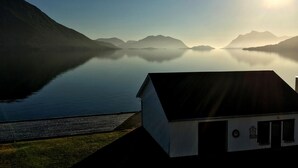 A serene lakeside view with a small building featuring a sloped roof and a door, surrounded by calm water and distant mountains.
