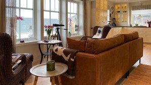 A cozy living room with a brown sofa, a wicker chair, and a small round table with a green vase. A telescope is on the windowsill, and a kitchen is visible in the background.