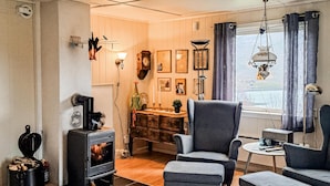 Ein gemütliches Wohnzimmer mit Kamin, einem bequemen Sessel, einem kleinen Tisch und Blick ins Freie durch ein Fenster.