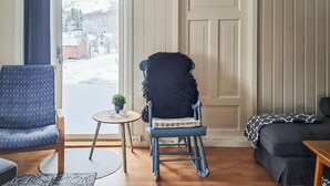 Ein gemütliches Wohnzimmer mit einem Schaukelstuhl, einem blauen Sessel und einer Couch. Ein kleiner Tisch mit Pflanze und Dekorationen ist zu sehen.