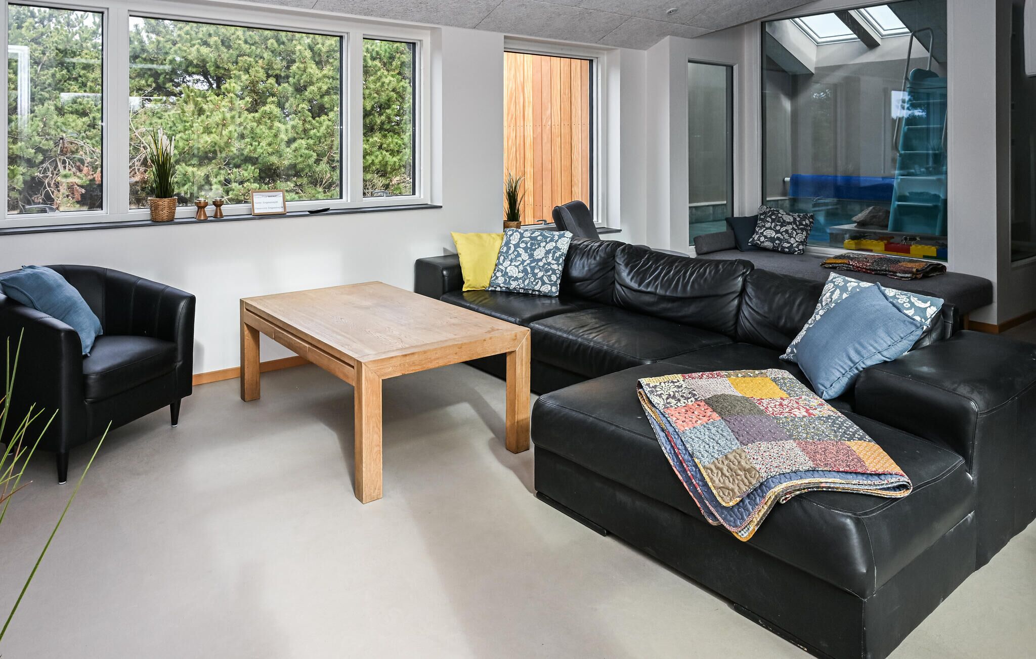 A modern living room with a black leather sofa, a wooden coffee table, and a view of trees through large windows.