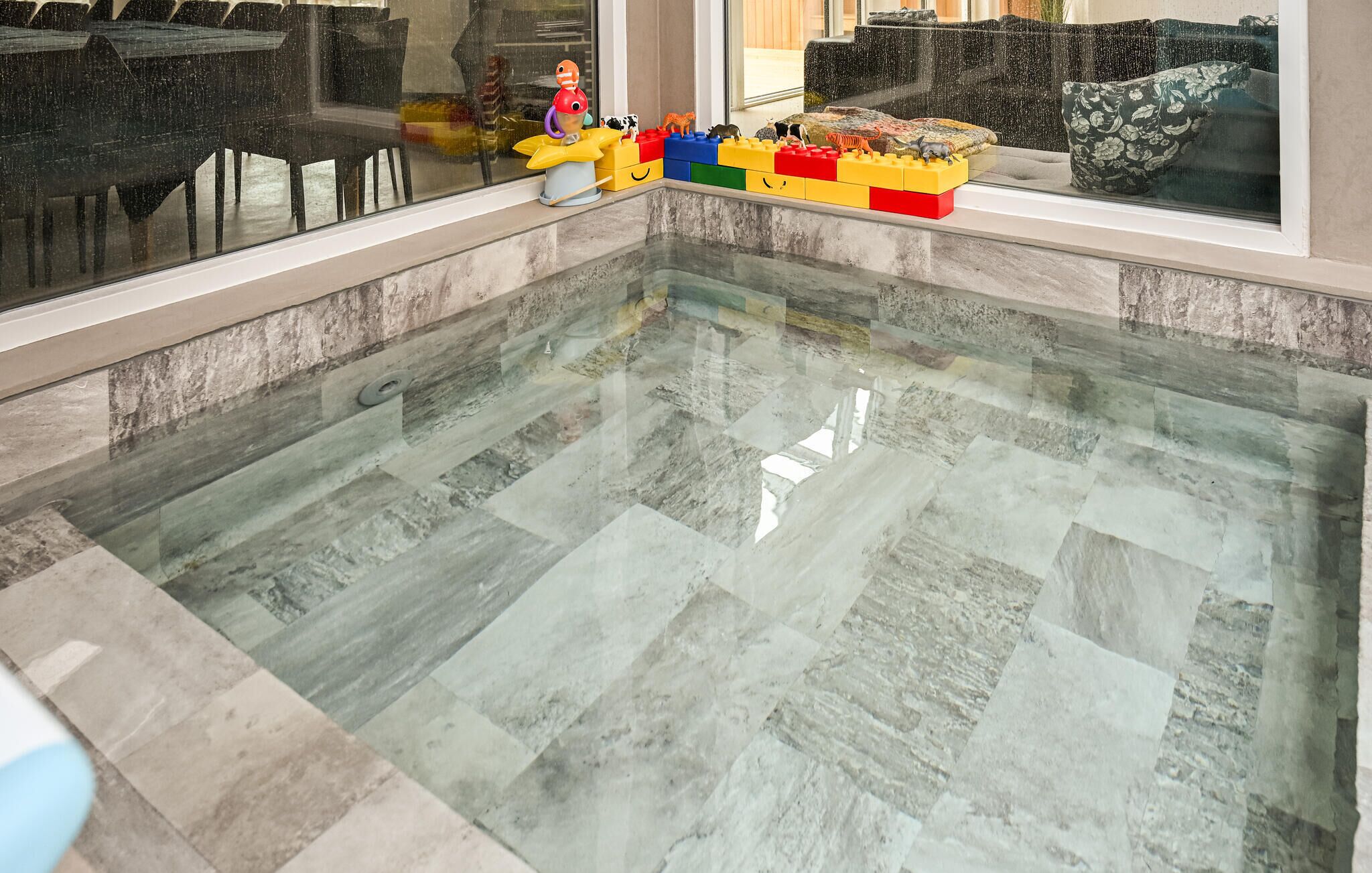Una piscina con fondo trasparente in vetro e pavimento piastrellato, circondata da una zona relax con divano e finestra.