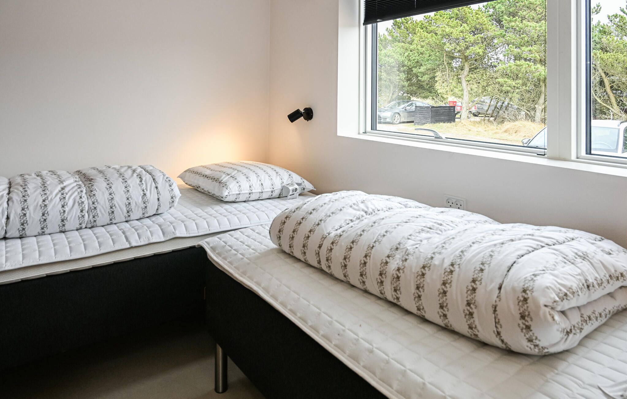 Ein Schlafzimmer mit zwei Betten, einem Fenster mit Blick auf Bäume und parkende Autos sowie einer Wandleuchte.