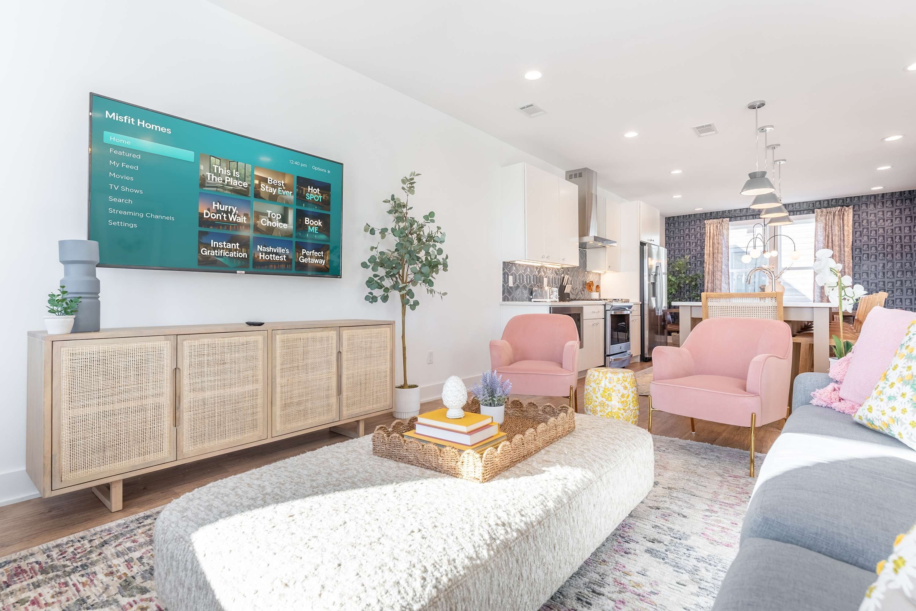 A modern living room with a large flat-screen TV displaying a streaming service interface, a rattan cabinet, a pink armchair, a grey sofa, and a decorative tray with books.