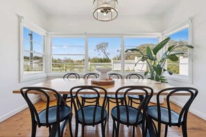 A dining room with a wooden table and black chairs, large windows with a view of greenery, and a hanging light fixture. - St Pauls Haven - 3brm Charming Cottage in Sorrento (Sorrento)