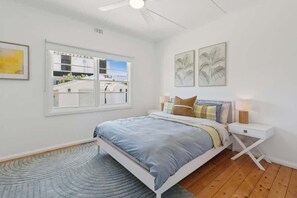 Ein Schlafzimmer mit Bett, Nachttischen, Deckenventilator und einem Fenster mit Blick auf Gebäude.
