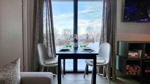 Un coin repas avec une table dressée pour quatre personnes, deux chaises de chaque côté, une grande fenêtre avec des rideaux et une cheminée sur laquelle est accrochée une télévision.