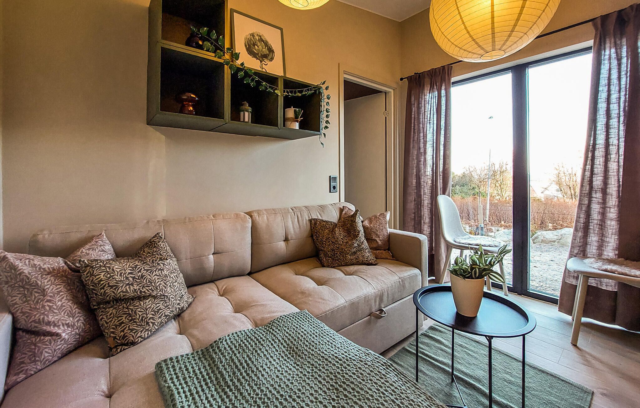 Sala de estar con un sofá color beige, cojines estampados, un plaid verde, una mesita redonda con una planta en maceta y un ventanal con cortinas.