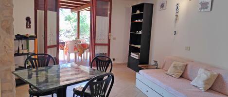 A dining area with a glass table and chairs, a sofa, a bookshelf, and a door leading outside.
