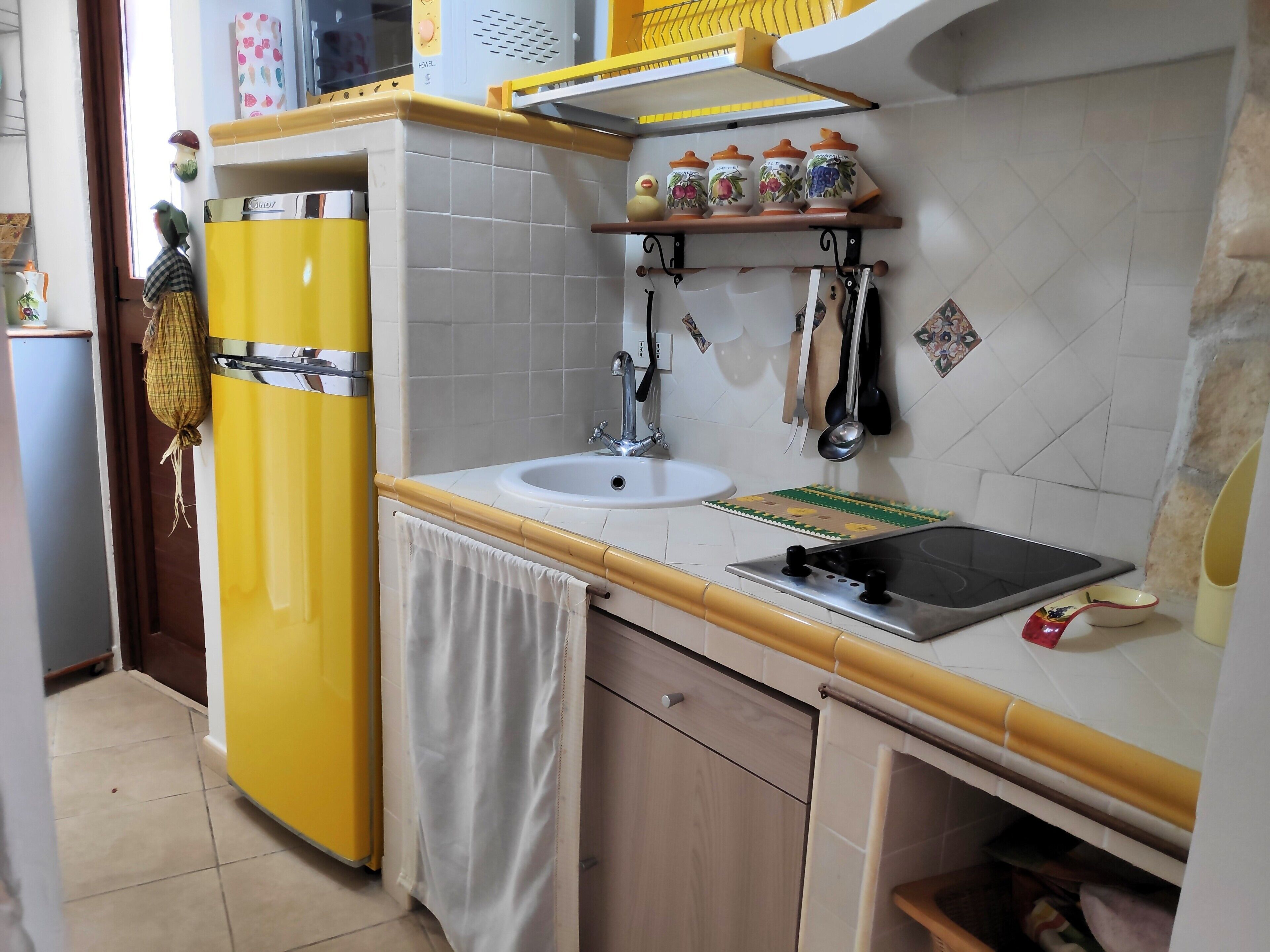 Een keuken met een gele koelkast, een spoelbak en een fornuis. Er staan decoratieve items op de muur en er is een raam boven de spoelbak.