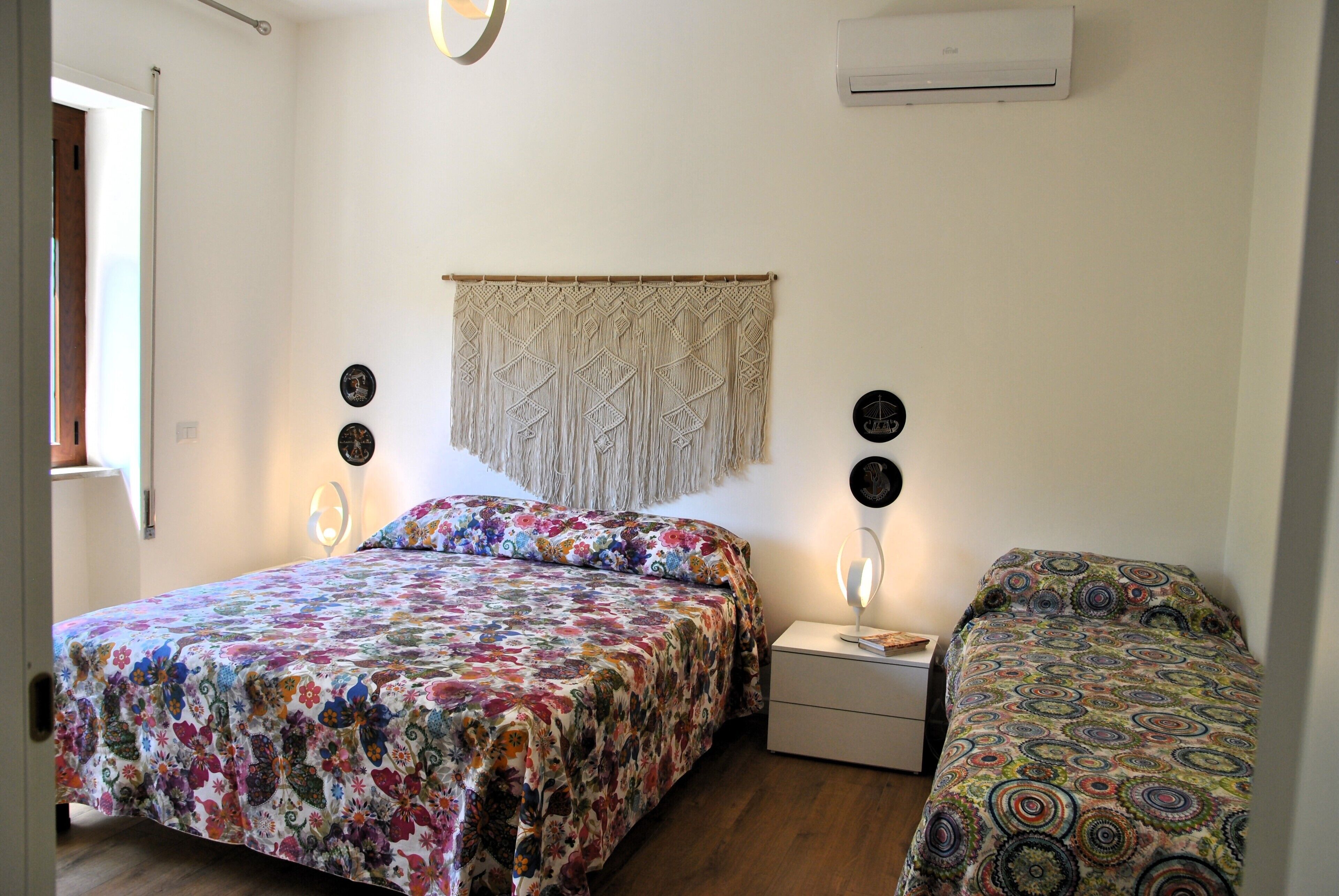 Un dormitorio con dos camas, un colgador de macramé en la pared y una mesita de noche con lámpara.
