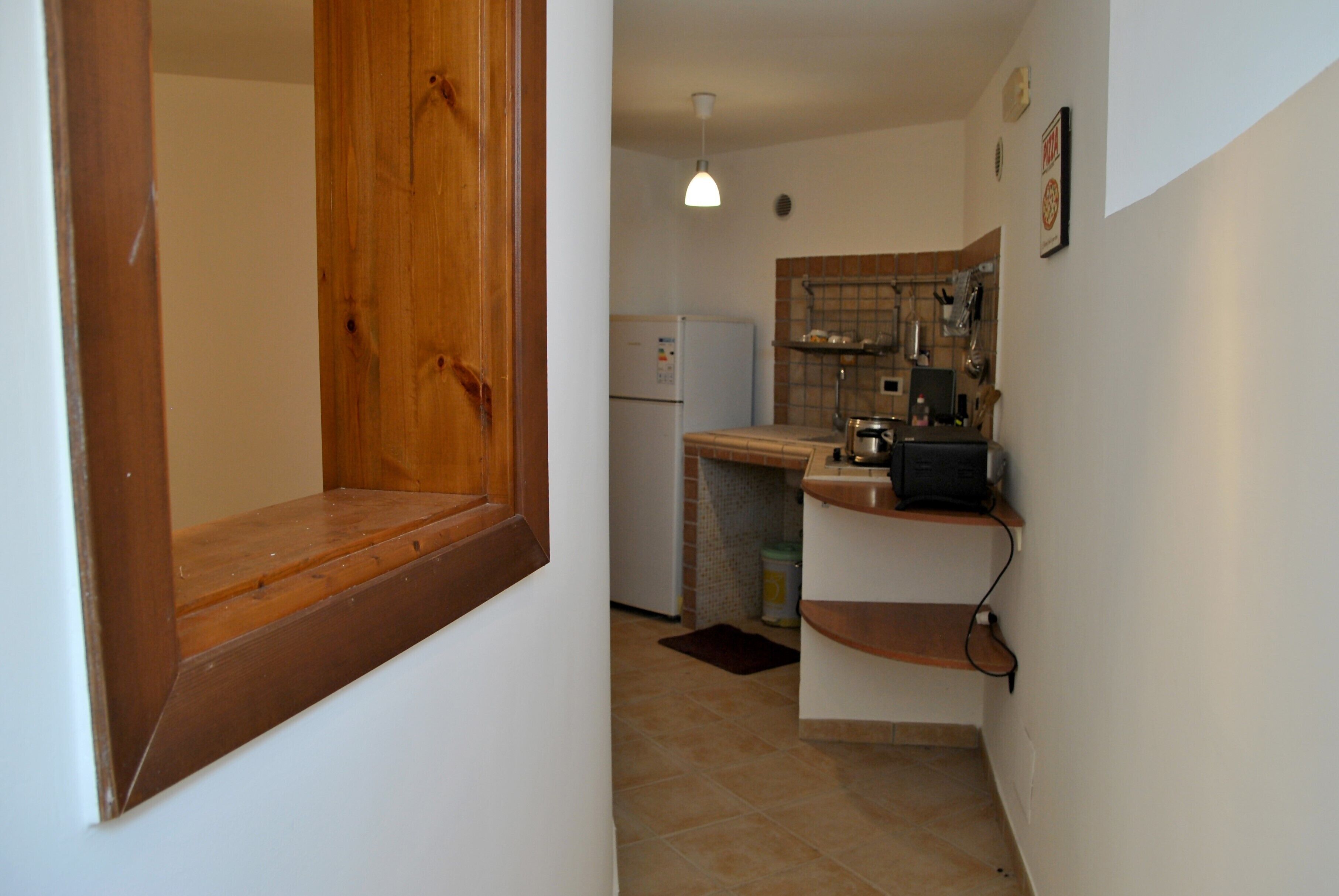 A narrow kitchen with a wooden door, a white refrigerator, and a small countertop with a sink and a kettle.