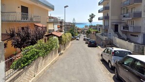 Una calle estrecha bordeada de edificios de varios pisos, algunos con balcones, coches estacionados y una vista despejada del mar en la distancia.